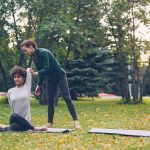 Diligent yoga student is learning Cow Face position sitting on mat and stretching arms while experienced teacher is helping her, girls are talking and laughing at outdoor ptactice.